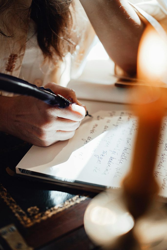 Crop unrecognizable female writer with feather taking notes in notepad at table on sunny day